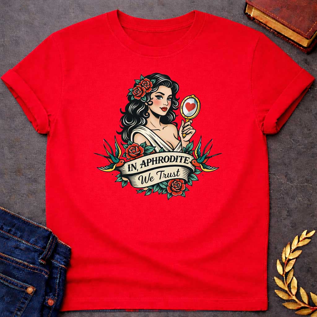 Red T-shirt graphic in a classic American traditional tattoo style featuring a pin-up illustration of Aphrodite, the Greek Goddess of Love, with dark wavy hair adorned with red roses, wearing a white draped gown, holding a gold hand mirror reflecting a red heart. She is flanked by two swooping swallows and surrounded by red roses and green leaves, with a scroll banner below reading "In Aphrodite We Trust" in bold and script lettering