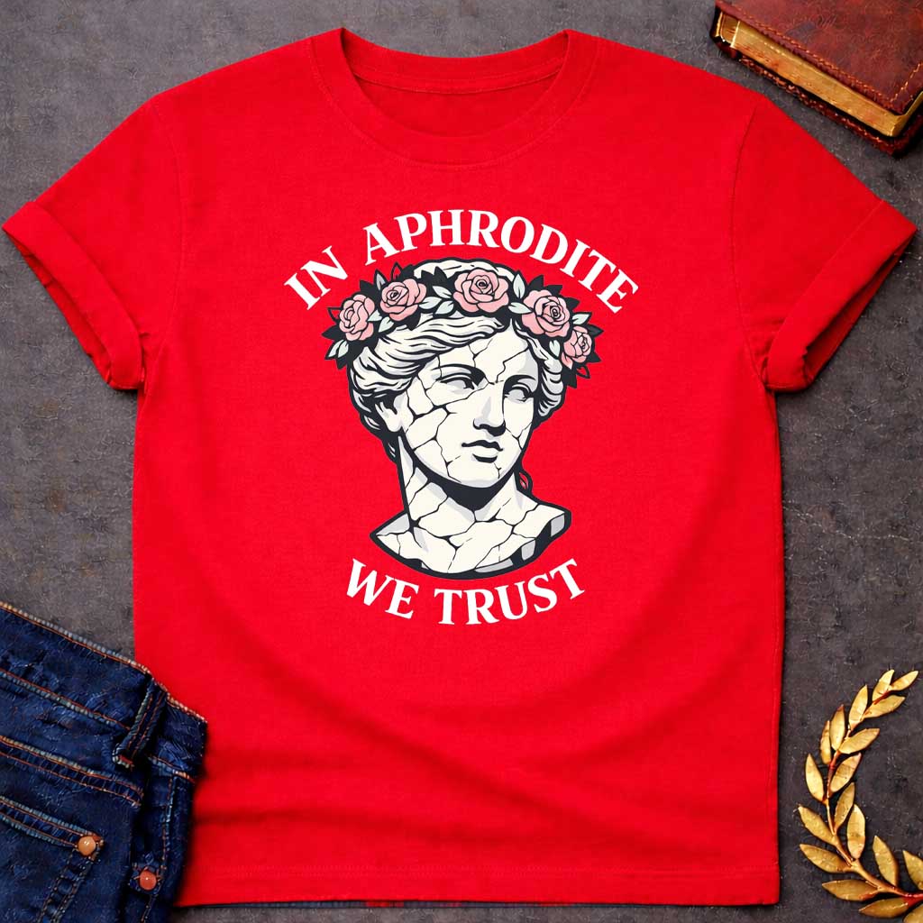 Red T-shirt graphic featuring a cracked marble bust illustration of Aphrodite, the Greek Goddess of Love, adorned with a floral crown of pink roses and green leaves, with bold arched text reading "In Aphrodite" curved above and "We Trust" curved below, in a modern aesthetic vaporwave-inspired style with white text and soft pink and white tones