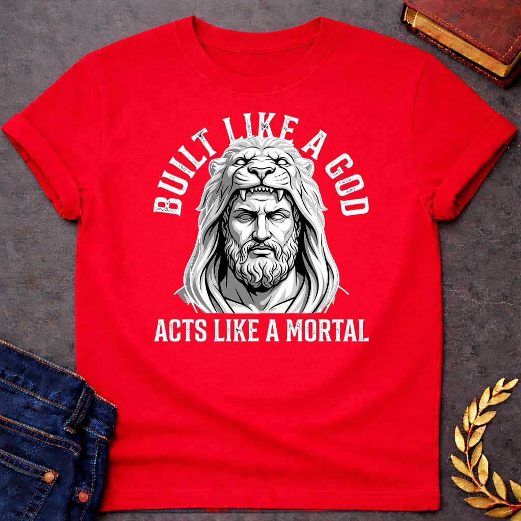 Red T-Shirt with a high-contrast, black-and-white graphic. The illustration depicts a stylized, statuesque man wearing the head and mane of a lion as a hood, reminiscent of Hercules. The text "BUILT LIKE A GOD" is arched over the top, and "ACTS LIKE A MORTAL" is printed in bold, straight text across the botto