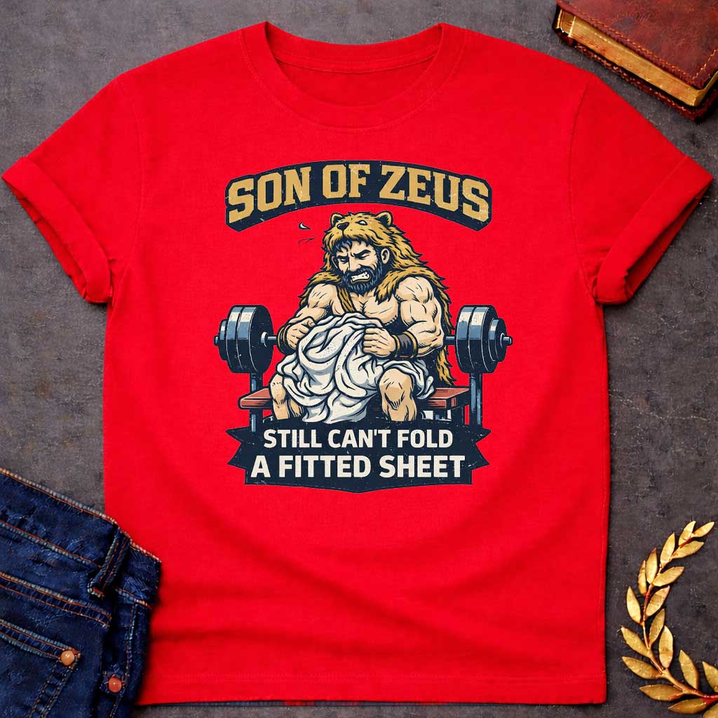 Red T-shirt featuring a humorous illustration of a muscular, bearded man wearing a lion pelt—resembling Hercules—sitting on a weight bench. He is struggling, with a frustrated expression, to fold a crumpled white fitted sheet. Behind him is a barbell. The text at the top reads "SON OF ZEUS" in a distressed, arched font, and the text at the bottom reads "STILL CAN'T FOLD A FITTED SHEET" in a banner