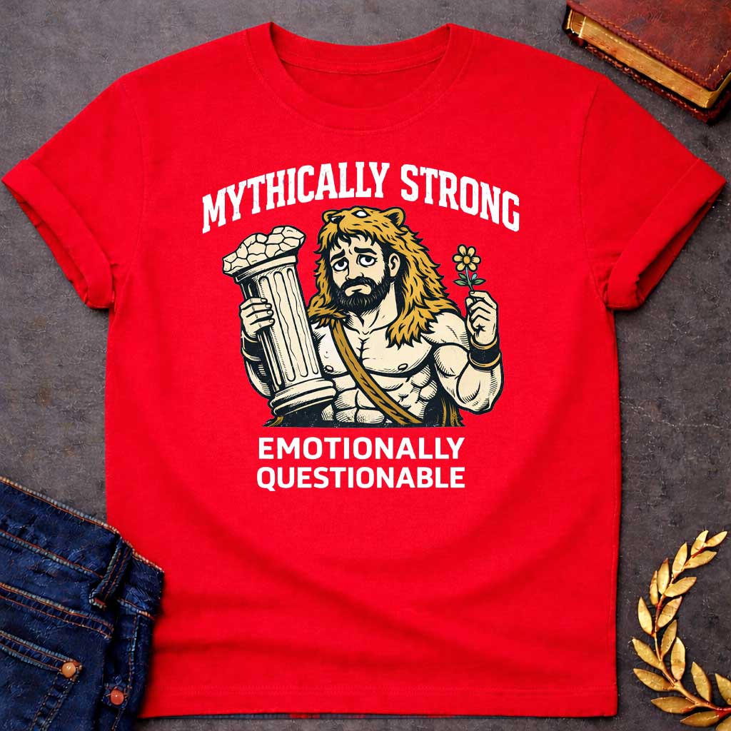 Red T-shirt featuring a humorous illustration of a muscular, bearded man wearing a lion-skin hood—reminiscent of Hercules—with a sad, weary expression. He holds a broken classical column in one hand and a small flower in the other. Arched over the top is the white text "MYTHICALLY STRONG," and below the illustration is the text "EMOTIONALLY QUESTIONABLE,"