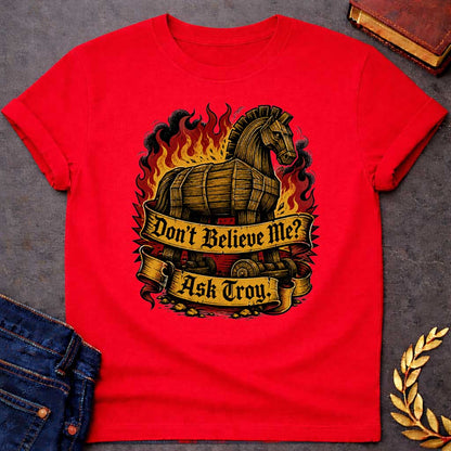 Red T-shirt with a bold illustration of the Trojan Horse engulfed in flames with red, orange, and yellow fire and black smoke. The wooden horse emerges from flames with detailed wood-grain texture. Two ornate gold banners with blackletter text read 'Don't Believe Me?' and 'Ask Troy.' Dark red background with rubble scattered below. References Cassandra's ignored warning about the Trojan Horse that led to Troy's destruction. Dramatic tattoo-inspired style with vintage engraving detail.