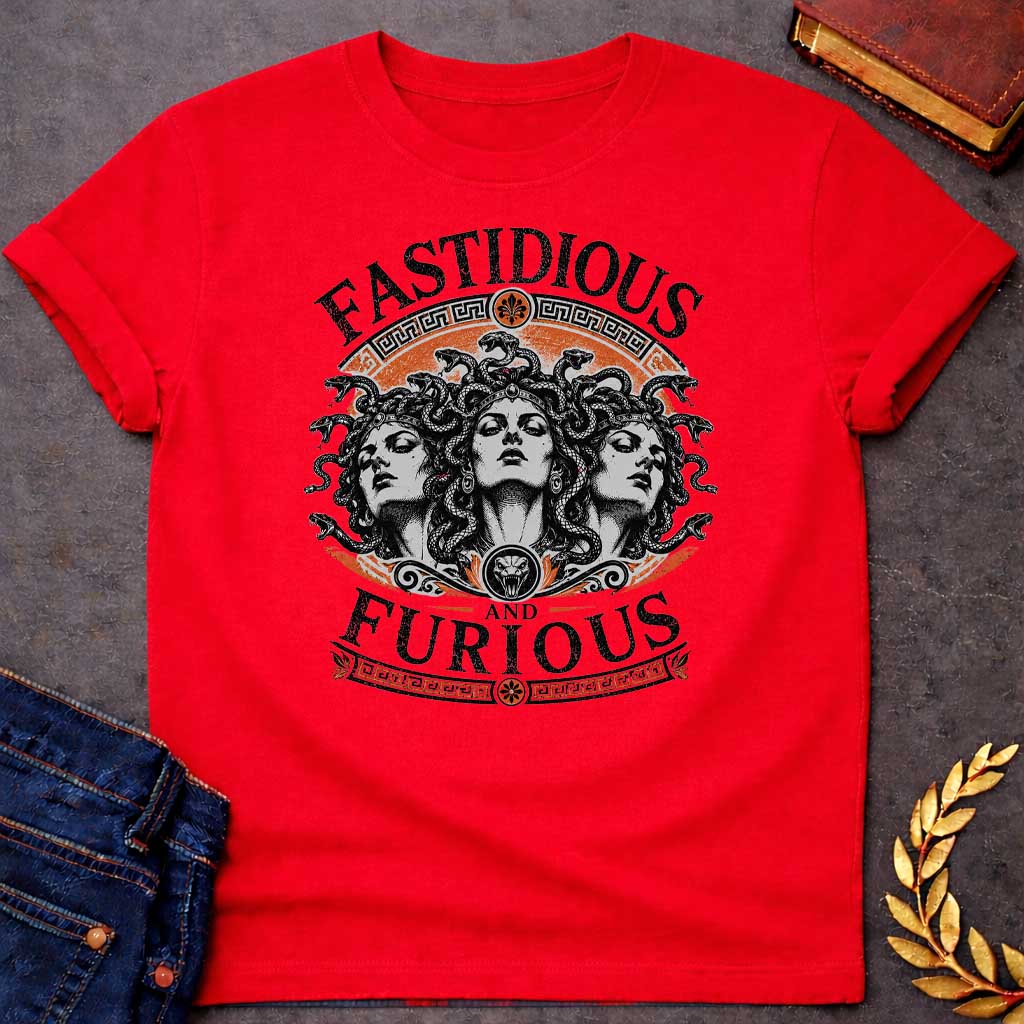 Red T-shirt with a Humorous design combining 'Fast and Furious' with Greek mythology. Text reads 'FASTIDIOUS AND FURIOUS' in distressed lettering surrounding three faces of Medusa in black and white engraving style. Detailed serpents with individual scales form her hair. Greek key pattern borders in orange frame the composition with decorative sun motifs and a small snake head icon at center. Blends classical mythology with contemporary pop culture humor and clever wordplay.