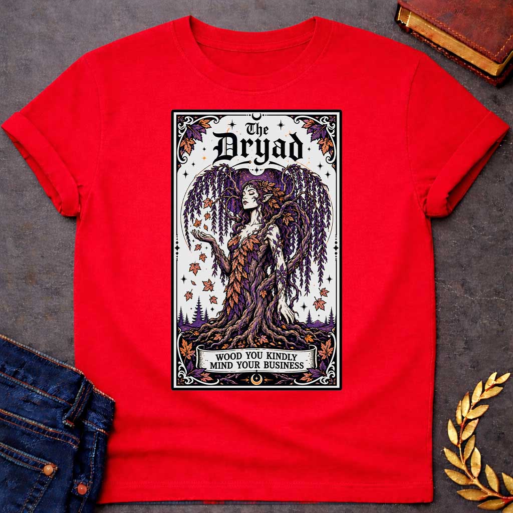 Red T-shirt with a mystical, tarot-card style design framed by an ornate border. It features a majestic weeping willow dryad whose gown is woven from purple and orange autumn leaves. At the top, bold gothic text reads "The Dryad." A weathered banner at the base of her rooted trunk features a clever, pun-based tagline that reads: "WOOD YOU KINDLY MIND YOUR BUSINESS" in a clean, black serif font