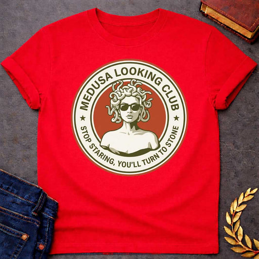Red t-shirt in retro badge style: a cool, sunglasses-wearing Medusa with snake hair stands confidently against a burnt-orange circle. Cream and olive-green ring text reads "Medusa Looking Club" above and "Stop Staring, You'll Turn to Stone" below, with two stars flanking the design.