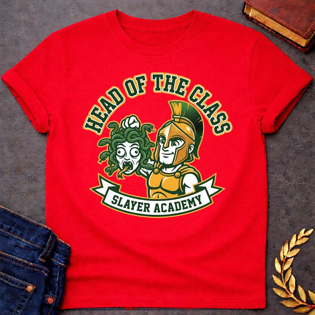 Red t-shirt in collegiate mascot style: a smirking gold-helmeted Greek warrior holds up Medusa's shocked, snarling severed head by her green snake hair. Bold green and gold text arcs above the design, with "Slayer Academy" on a white ribbon banner below. Playful varsity-logo aesthetic on a white background.