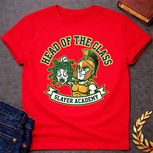 Red t-shirt in collegiate mascot style: a smirking gold-helmeted Greek warrior holds up Medusa's shocked, snarling severed head by her green snake hair. Bold green and gold text arcs above the design, with "Slayer Academy" on a white ribbon banner below. Playful varsity-logo aesthetic on a white background.