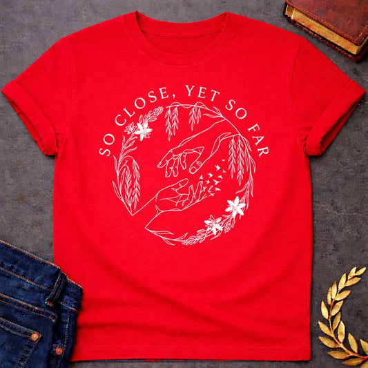 Red t-shirt in delicate fine-line style: two reaching hands almost touch, with one hand dissolving into a flock of small birds mid-fingertip, evoking Orpheus and Eurydice. Willow branches and small flowers form a circular wreath around the scene, with elegant serif text arcing above