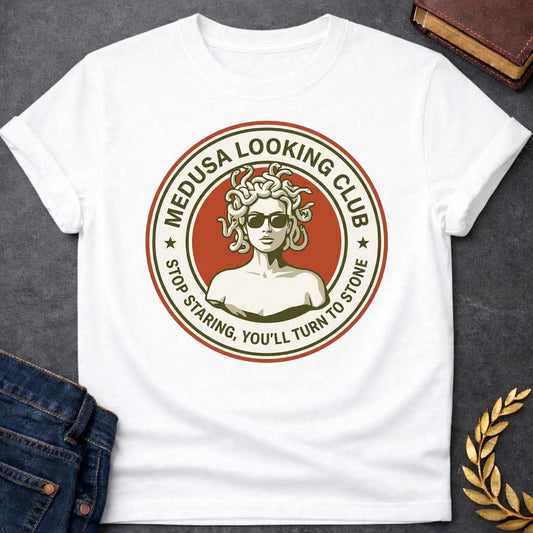 White t-shirt in retro badge style: a cool, sunglasses-wearing Medusa with snake hair stands confidently against a burnt-orange circle. Cream and olive-green ring text reads "Medusa Looking Club" above and "Stop Staring, You'll Turn to Stone" below, with two stars flanking the design.