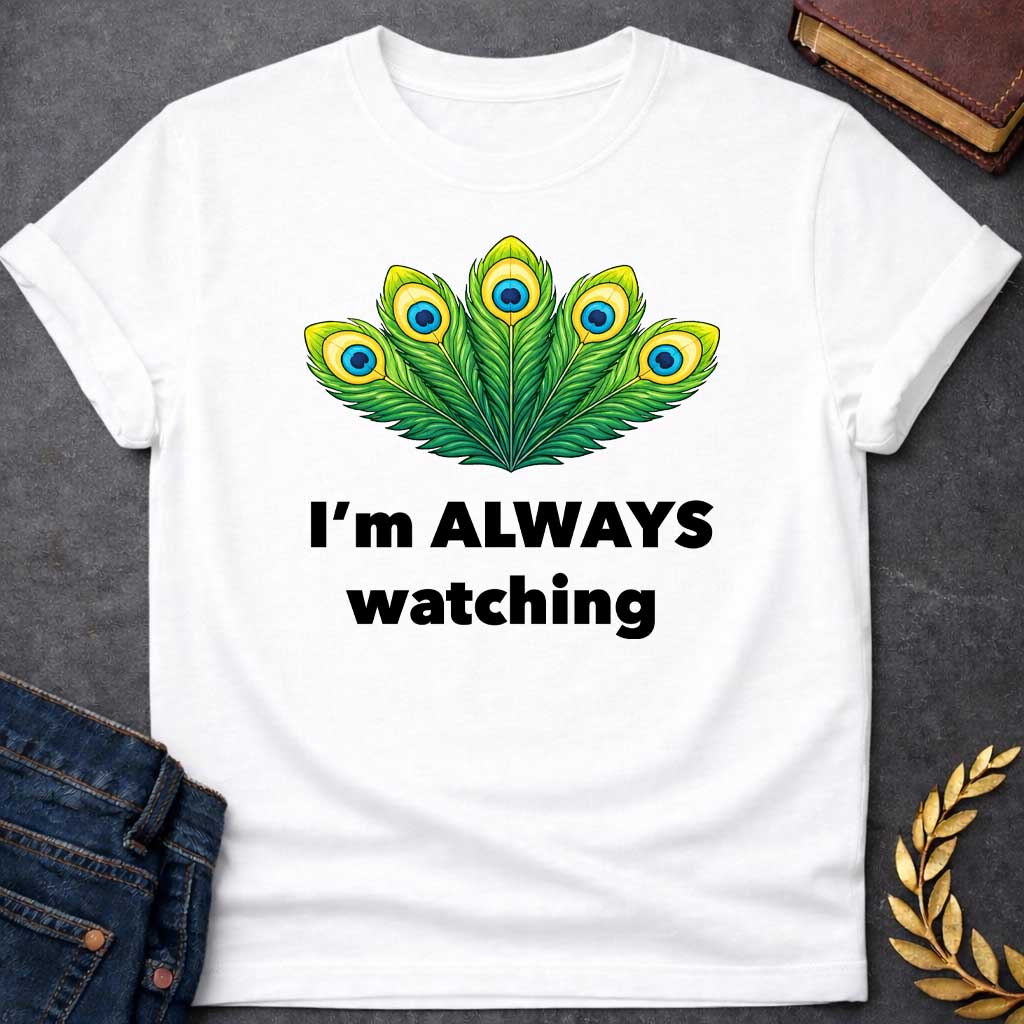 White T-shirt graphic featuring a vibrant illustrated fan of five peacock feathers in rich green, yellow, and blue tones, each with a distinctive eye-spot, above text reading "I'm Always Watching" — a humorous reference to Hera, the Greek Queen of the Gods, whose sacred symbol is the peacock