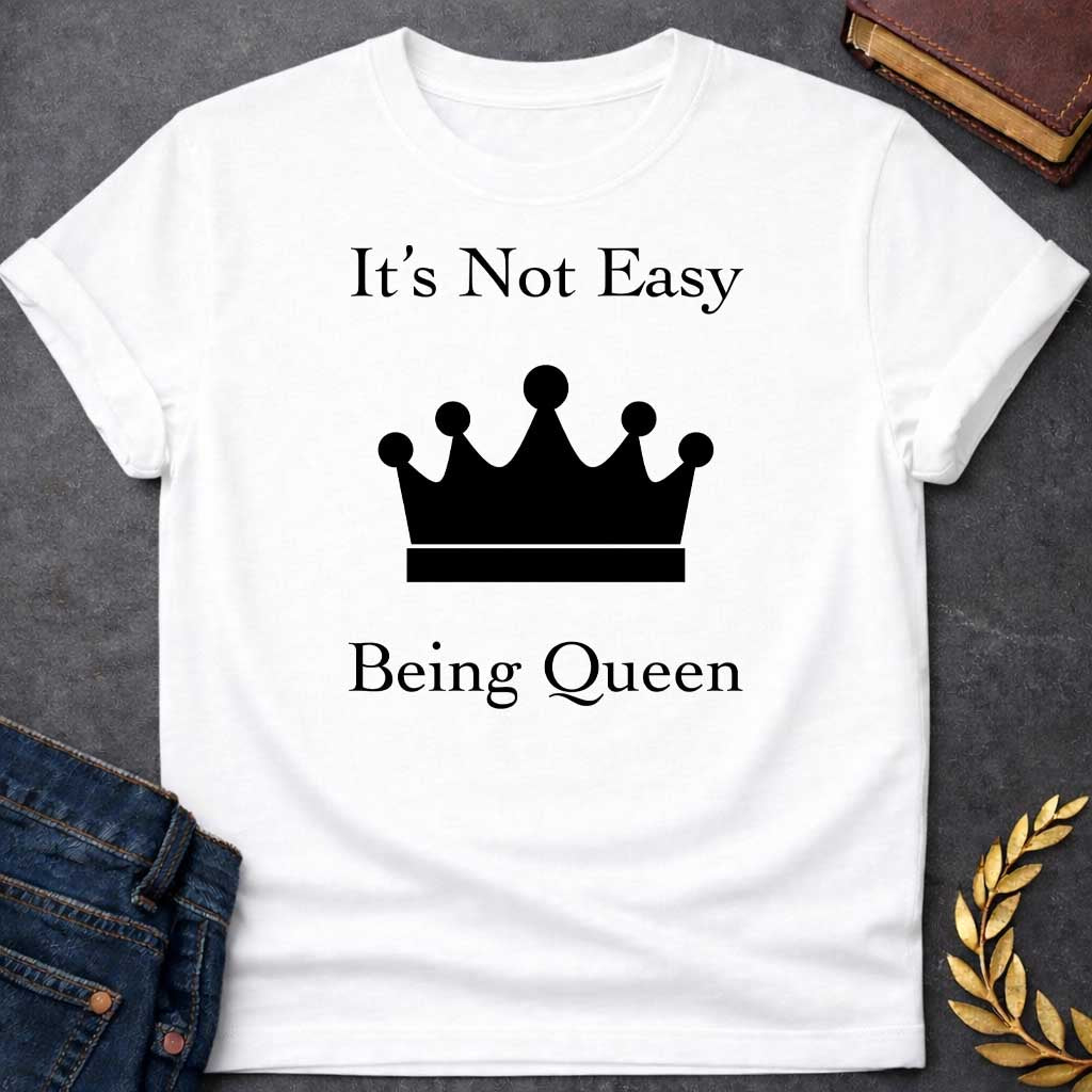 White T-shirt graphic in white on black featuring elegant serif text reading "It's Not Easy Being Queen" split above and below a simple white crown silhouette icon, in a humorous nod to Hera, the Greek Queen of the Gods