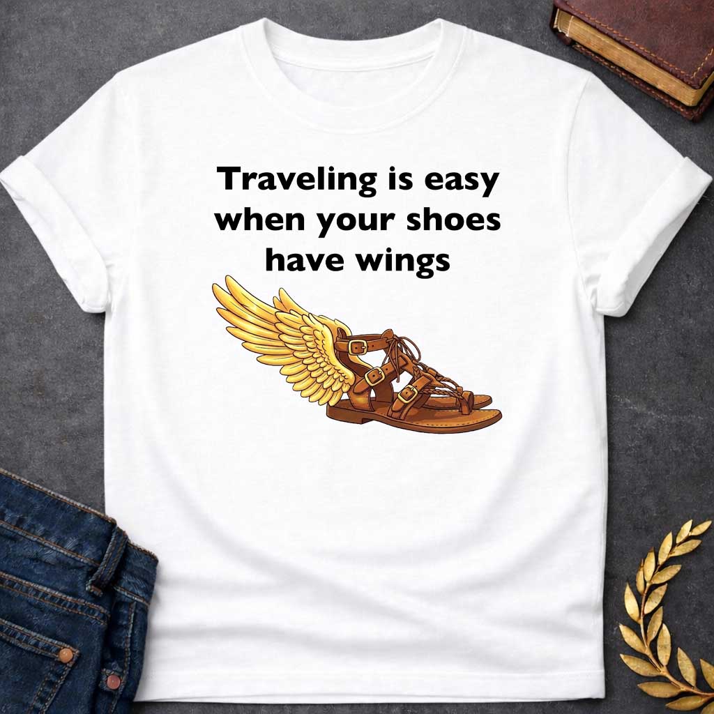 White T-shirt graphic featuring bold black text reading "Traveling is easy when your shoes have wings" above a detailed illustration of Hermes' winged sandal, depicted as a brown leather gladiator sandal with gold buckles and braided lacing, with a large golden wing extending from the ankle — a humorous reference to the talaria, the iconic flying sandals of Hermes, the Greek Messenger God
