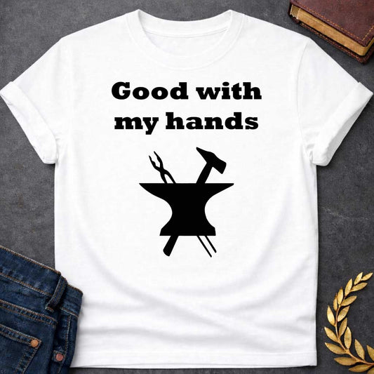 White T-shirt graphic in black featuring bold serif text reading "Good with my hands" above a flat black silhouette illustration of a blacksmith's anvil with a hammer and tongs crossed on top — a humorous nod to Hephaestus, the Greek God of the Forge