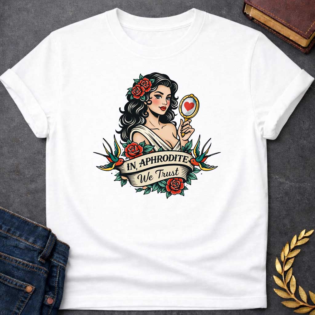 White T-shirt graphic in a classic American traditional tattoo style featuring a pin-up illustration of Aphrodite, the Greek Goddess of Love, with dark wavy hair adorned with red roses, wearing a white draped gown, holding a gold hand mirror reflecting a red heart. She is flanked by two swooping swallows and surrounded by red roses and green leaves, with a scroll banner below reading "In Aphrodite We Trust" in bold and script lettering