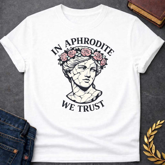 White T-shirt graphic featuring a cracked marble bust illustration of Aphrodite, the Greek Goddess of Love, adorned with a floral crown of pink roses and green leaves, with bold arched text reading "In Aphrodite" curved above and "We Trust" curved below, in a modern aesthetic vaporwave-inspired style with white text and soft pink and white tones