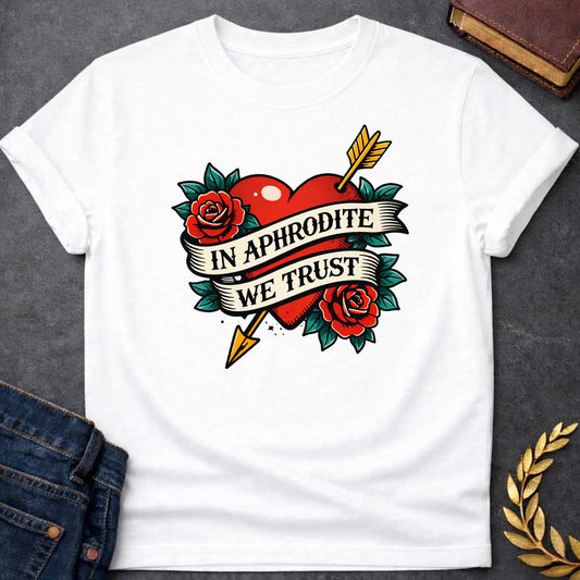 White T-shirt graphic in a classic American traditional tattoo style featuring a bold red heart pierced diagonally by a gold arrow, flanked by two red roses with teal green leaves, with a cream scroll banner wrapped around the heart reading "In Aphrodite We Trust" in black serif text — referencing Aphrodite, the Greek Goddess of Love, and Eros whose golden arrow inspired love