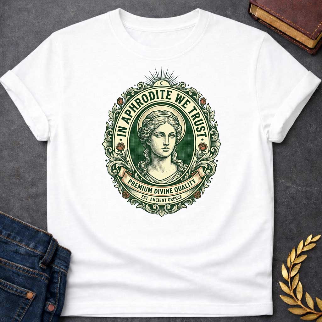 White T-shirt graphic featuring a vintage apothecary label-style oval badge design in forest green, cream, and terracotta. A detailed engraving-style portrait of Aphrodite is centered within the badge, surrounded by ornate scrollwork and flanked by red roses, with a crescent moon and sunburst radiating from the top. Text arched around the inner circle reads "In Aphrodite We Trust," with a scroll banner below reading "Premium Divine Quality" and smaller text underneath reading "Est. Ancient Greece,"