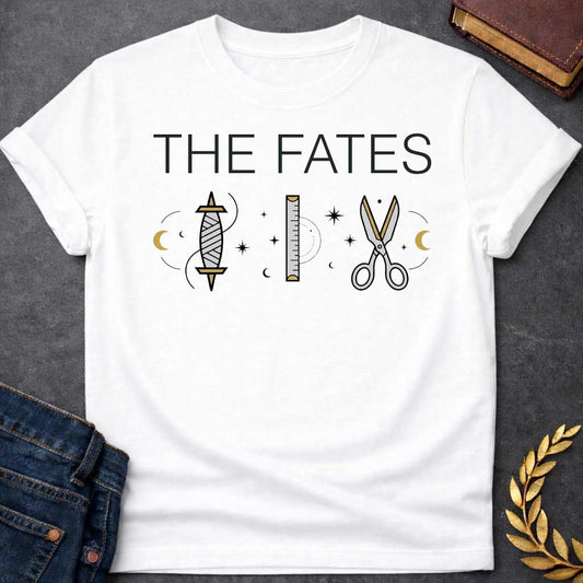 White T-shirt graphic featuring large bold black text reading "The Fates" above three minimalist icons representing the tools of the Moirai, the Greek Fates — a spindle with thread, a measuring ruler, and a pair of scissors — each surrounded by subtle crescent moons and celestial star details, rendered in white and gold tones