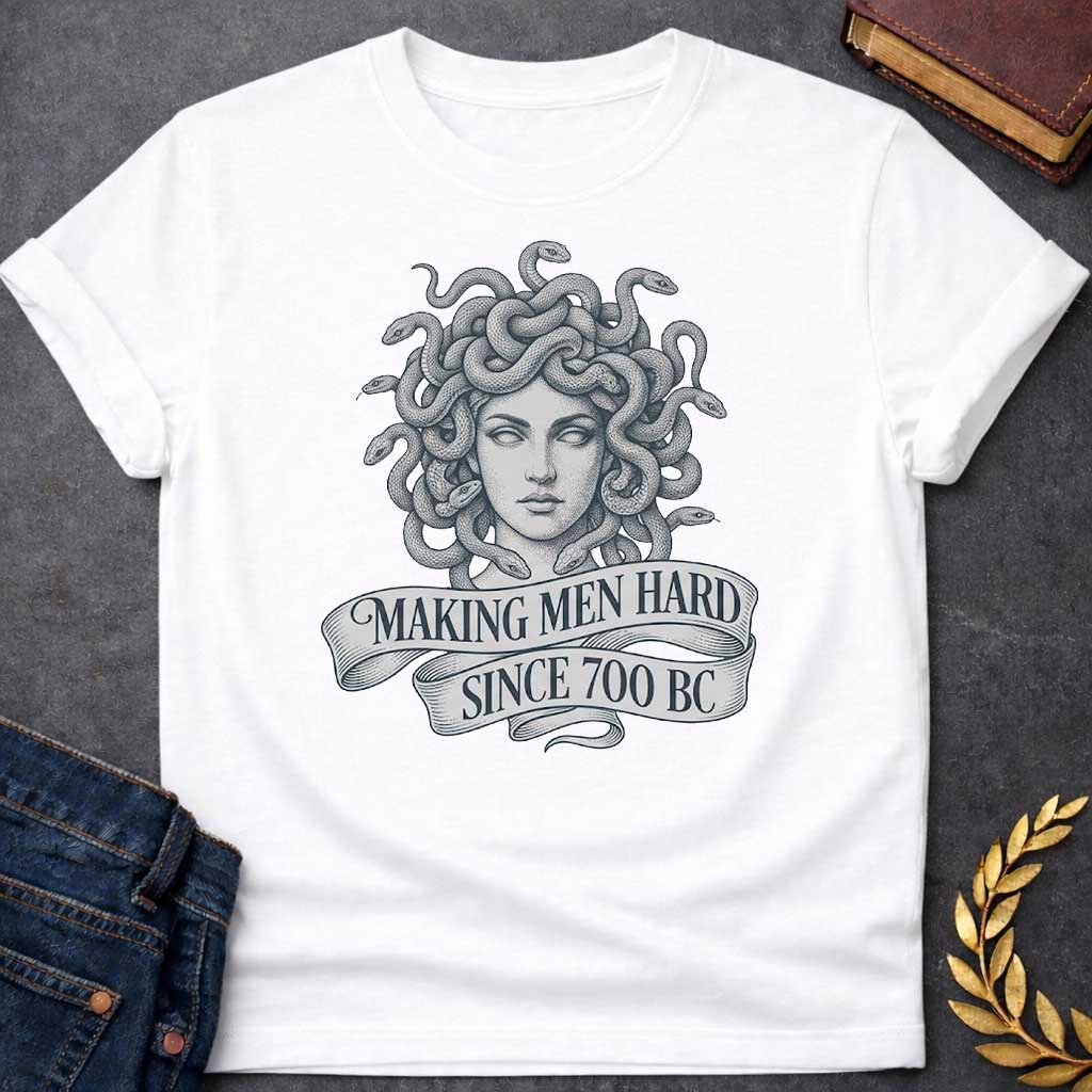White T-shirt graphic in grey and white featuring a highly detailed fine-line tattoo-style front-facing portrait of Medusa with numerous coiling snakes for hair, rendered in intricate stippling and engraving technique, with a flowing ribbon banner below reading "Making Men Hard Since 700 BC" — a witty double entendre referencing Medusa's mythological power to turn men to stone