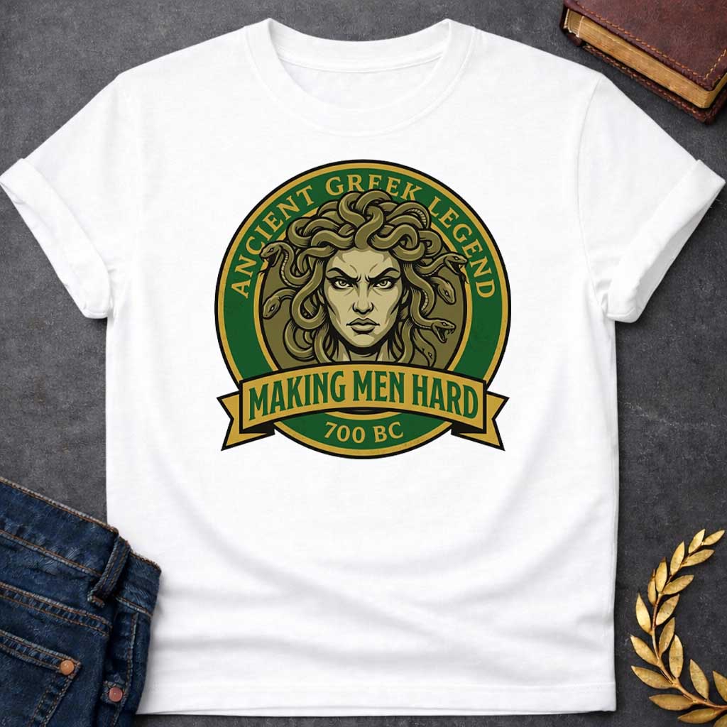 White T-shirt graphic featuring a bold vintage badge design in forest green and gold. A fierce front-facing illustration of Medusa with coiling olive-toned snakes for hair and an intense glaring expression fills the circular center, with gold arched text reading "Ancient Greek Legend" around the upper border. A gold ribbon banner below the portrait reads "Making Men Hard" in green text, with "700 BC" below it — a witty double entendre referencing Medusa's mythological power to turn men to stone