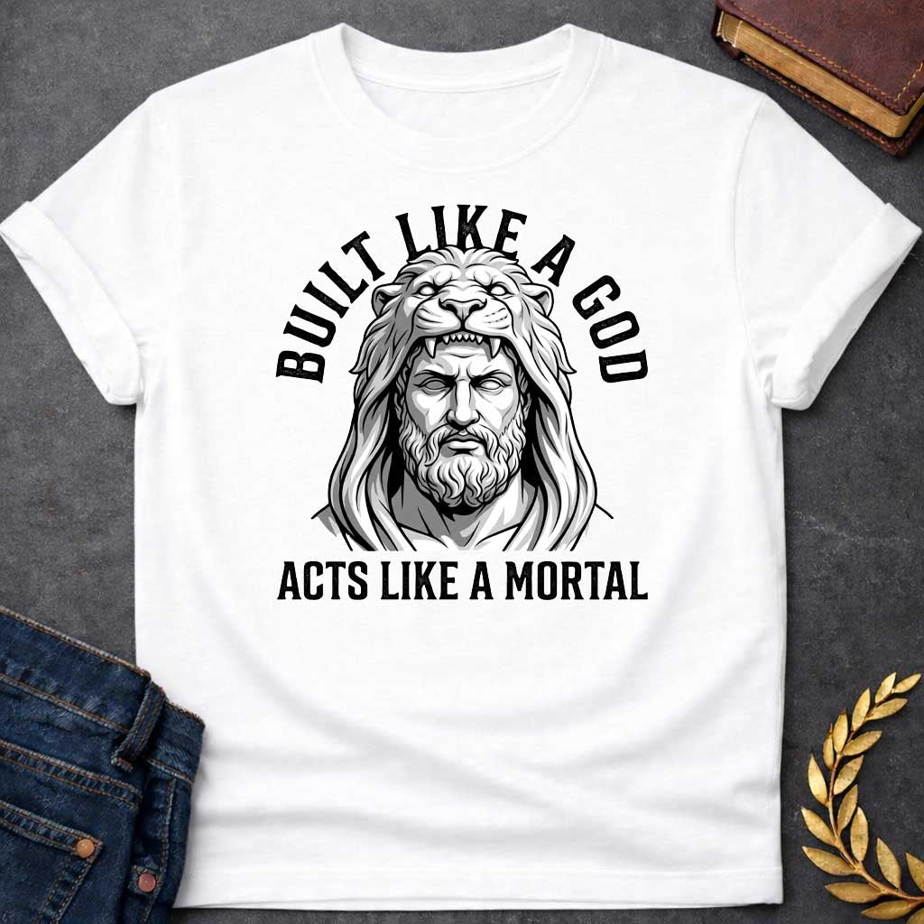 White T-Shirt with a high-contrast, black-and-white graphic. The illustration depicts a stylized, statuesque man wearing the head and mane of a lion as a hood, reminiscent of Hercules. The text "BUILT LIKE A GOD" is arched over the top, and "ACTS LIKE A MORTAL" is printed in bold, straight text across the botto