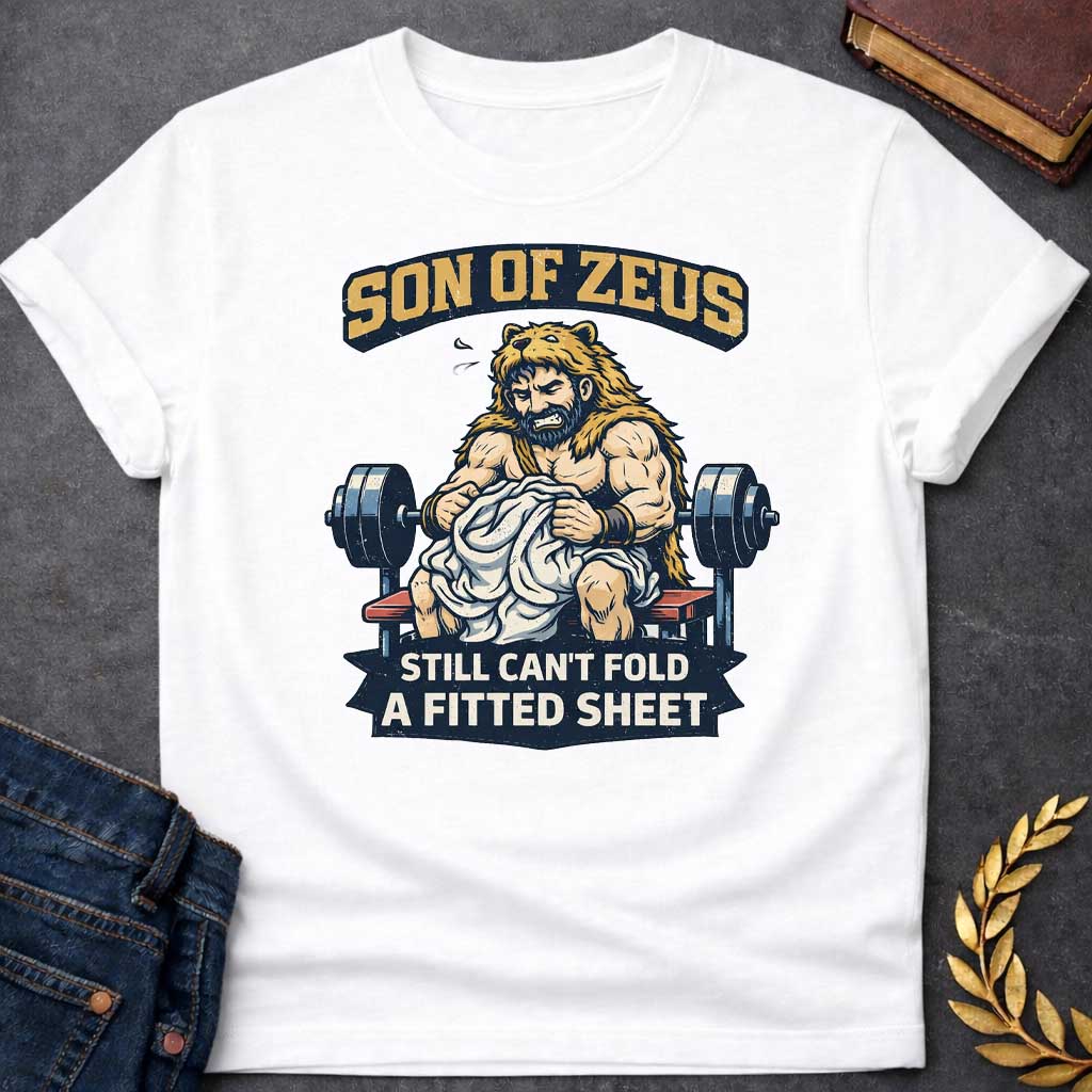 White T-shirt featuring a humorous illustration of a muscular, bearded man wearing a lion pelt—resembling Hercules—sitting on a weight bench. He is struggling, with a frustrated expression, to fold a crumpled white fitted sheet. Behind him is a barbell. The text at the top reads "SON OF ZEUS" in a distressed, arched font, and the text at the bottom reads "STILL CAN'T FOLD A FITTED SHEET" in a banner
