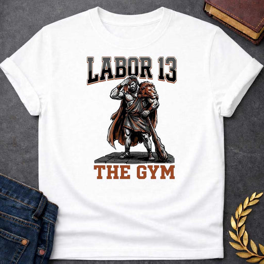 White T-shirt featuring a sweat-drenched, muscular figure resembling Heracles, wearing a lion-skin cloak and holding a large wooden club. He stands on a stone platform while wiping his brow with a towel. The text "LABOR 13" is printed in large, bold, block letters across the top, and "THE GYM" is in a matching style at the bottom