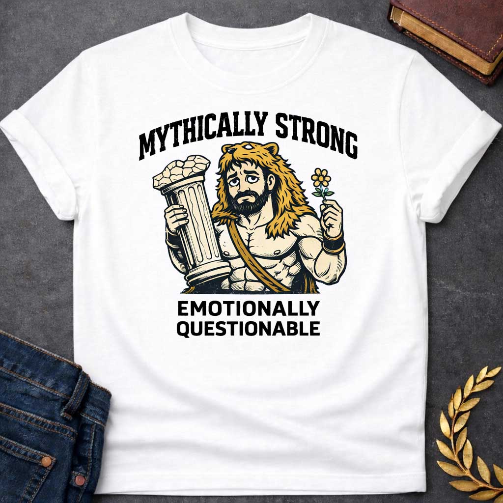 White T-shirt featuring a humorous illustration of a muscular, bearded man wearing a lion-skin hood—reminiscent of Hercules—with a sad, weary expression. He holds a broken classical column in one hand and a small flower in the other. Arched over the top is the black text "MYTHICALLY STRONG," and below the illustration is the text "EMOTIONALLY QUESTIONABLE,"