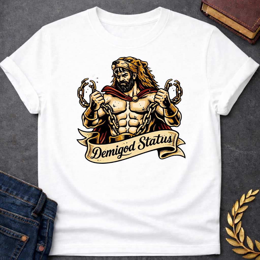 White T-shirt featuring a muscular, bearded figure resembling Heracles, wearing a lion-skin hood and a red cape, as he heroically snaps thick golden chains with his hands. Below the illustration is a flowing, decorative banner with the text "Demigod Status" written in a stylish script font,