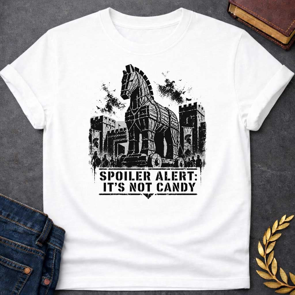 White T-shirt with a graphic in black featuring a gritty detailed ink splatter-style illustration of the Trojan Horse towering over the walls of Troy, flanked by Spartan warriors with shields and spears and castle battlements with birds circling overhead. Bold military stencil text in a distressed banner below reads "Spoiler Alert: It's Not Candy" — a humorous reference to the Greek legend of the Trojan War