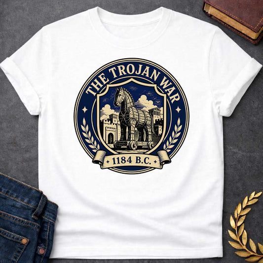 White T-shirt with a graphic featuring a formal vintage seal design in navy and cream. A detailed engraving-style illustration of the Trojan Horse on wheels is centered within a shield, set against the walled city of Troy with Greek temple columns and dramatic clouds. Text arched around the outer circle reads "The Trojan War" with gold diamond stars, flanked by cream laurel branches, and a scroll banner below reads "1184 B.C." — referencing the legendary Trojan War from Greek mythology