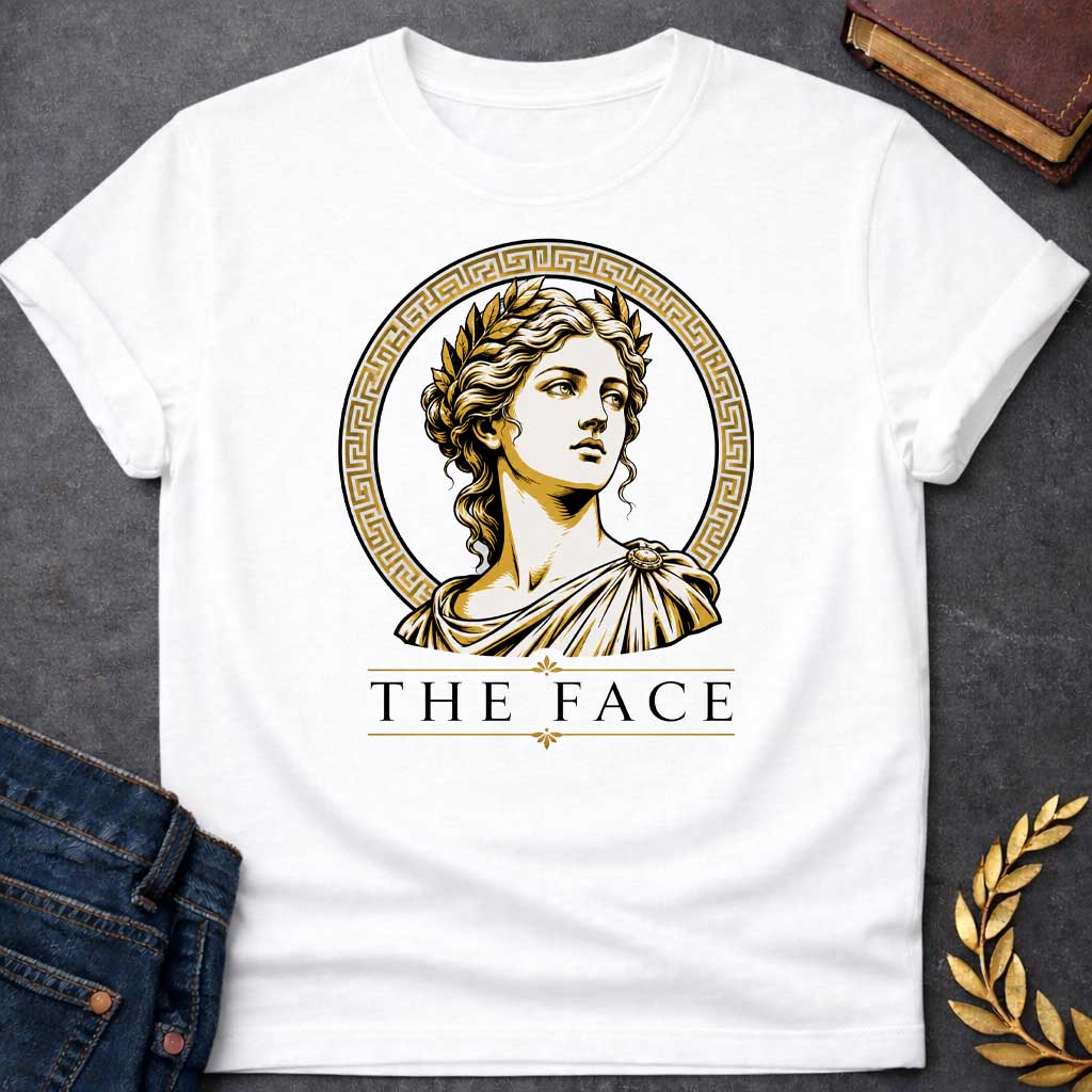White T-shirt with a classical Greek graphic featuring a gold and cream vector portrait of Helen of Troy in profile. She wears a golden laurel wreath and a traditional draped chiton, looking upward with a serene expression. The portrait is framed by a circular border with a gold Greek key (meander) pattern. Below the emblem, the text "THE FACE" is written in a clean, elegant black serif font between horizontal gold accent lines.
