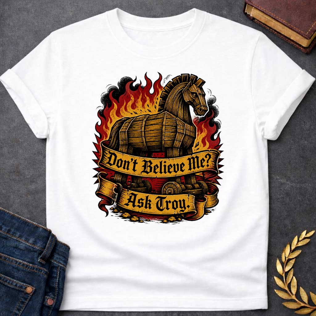 White T-shirt with a bold illustration of the Trojan Horse engulfed in flames with red, orange, and yellow fire and black smoke. The wooden horse emerges from flames with detailed wood-grain texture. Two ornate gold banners with blackletter text read 'Don't Believe Me?' and 'Ask Troy.' Dark red background with rubble scattered below. References Cassandra's ignored warning about the Trojan Horse that led to Troy's destruction. Dramatic tattoo-inspired style with vintage engraving detail.