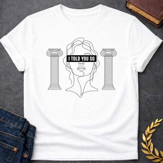 White T-shirt with a minimalist line art design featuring a continuous black ink sketch of Cassandra or a Greek goddess. Her eyes are covered by a bold, solid black rectangular bar with the text "I TOLD YOU SO" in white sans-serif capital letters. The portrait is flanked on both sides by simple, symmetrical line-drawn Ionic Greek columns.