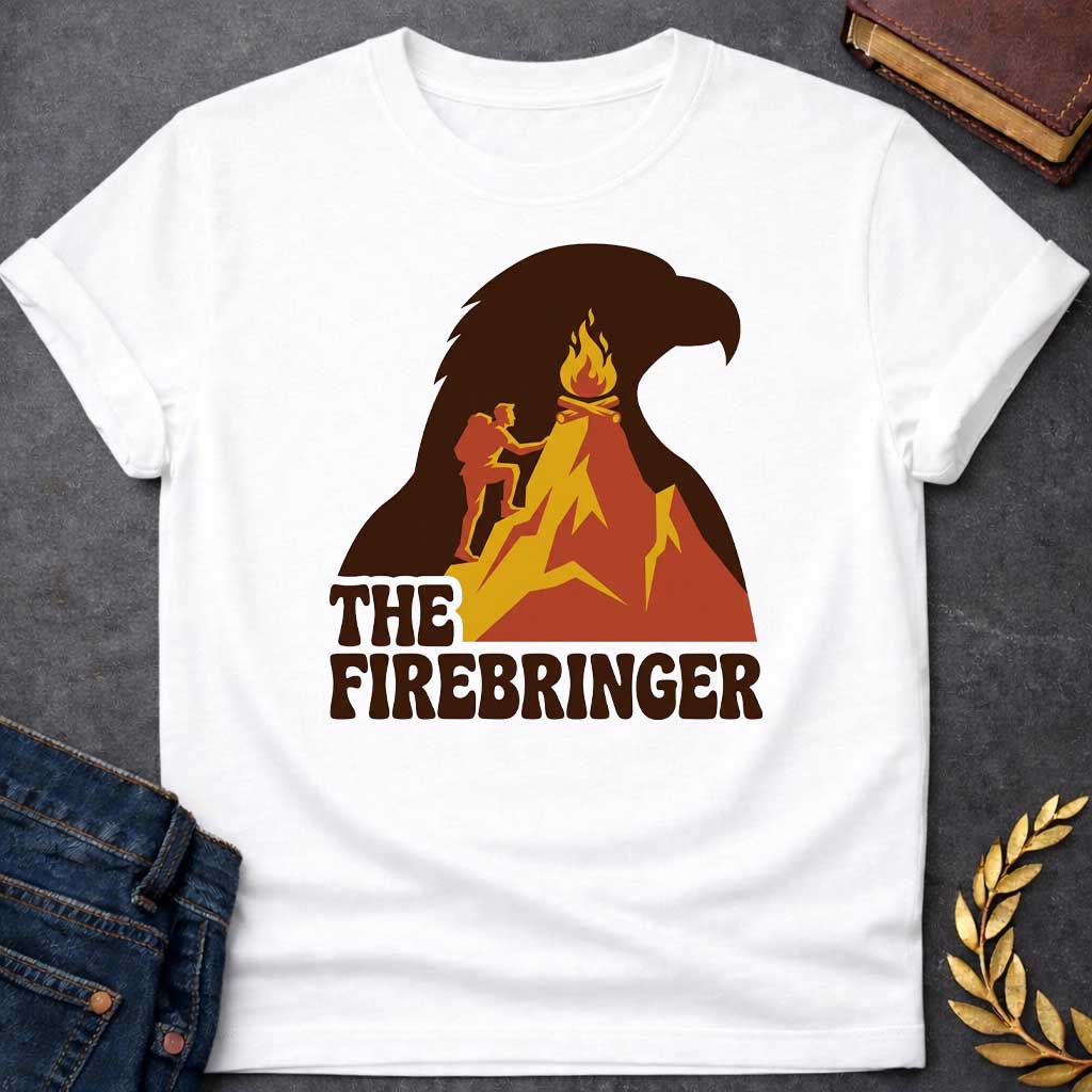 White T-shirt with a retro, outdoor-style t-shirt graphic referencing Prometheus. It features a silhouette of a hiker with a backpack climbing an orange and yellow mountain peak toward a campfire at the top. The entire mountain scene is set against a large background silhouette of an eagle's head in dark brown. Below the image, the text "THE FIREBRINGER" is written in a bold, stylized 1970s-style brown font.
