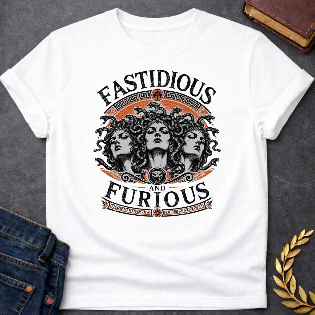 White T-shirt with a Humorous design combining 'Fast and Furious' with Greek mythology. Text reads 'FASTIDIOUS AND FURIOUS' in distressed lettering surrounding three faces of Medusa in black and white engraving style. Detailed serpents with individual scales form her hair. Greek key pattern borders in orange frame the composition with decorative sun motifs and a small snake head icon at center. Blends classical mythology with contemporary pop culture humor and clever wordplay.