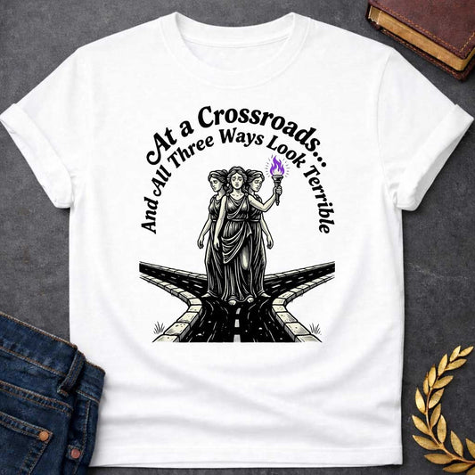 White T-shirt with an Illustration of the Hecate at a crossroads, rendered in black and white engraving style. The figures wear flowing robes and stand together, with the center holding a flaming torch with purple flames. Text reads 'At a Crossroads: And All Three Ways Look Terrible,' humorously referencing difficult choices and Hecate’s role as goddess of the crossroads. Multiple roads branch beneath them in perspective, receding into darkness symbolizing uncertainty.