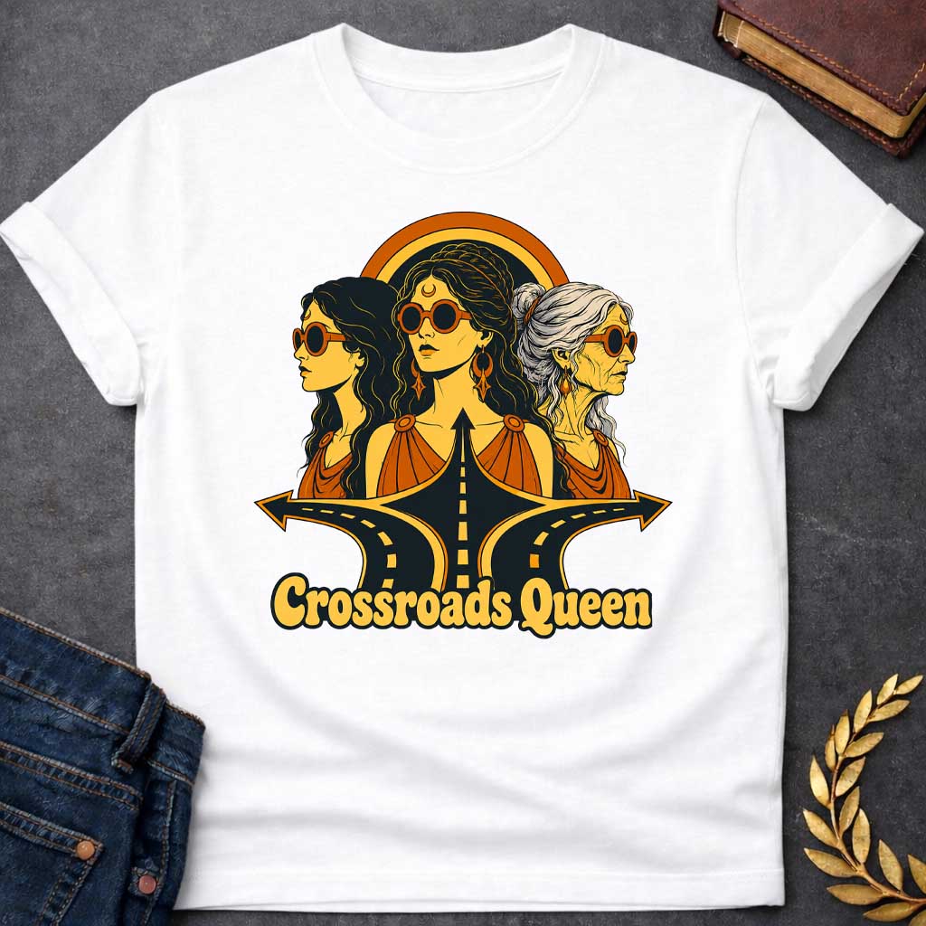 White T-shirt with a Glamorous retro illustration of Hecate as 'Crossroads Queen' in gold and orange with 70s aesthetic. Three female faces wear oversized round sunglasses and gold jewelry including crescent moon symbols and hoops. Center figure wears a crescent moon bindi. All wear draped orange clothing. A golden halo radiates behind them. Below, stylized black roads with yellow center lines converge at a crossroads. Text reads 'Crossroads Queen' in gold.