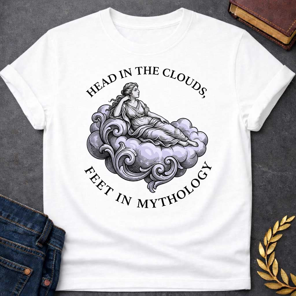 White T-shirt with a classical Greek mythology t-shirt design featuring a detailed engraving-style illustration. A woman in a traditional draped chiton relaxes gracefully, reclining on a large, swirling baroque-style cloud shaded in a pale purple hue. Arched above her is the text "HEAD IN THE CLOUDS," and curved inversely below the cloud is "FEET IN MYTHOLOGY," all written in a clean black serif font.