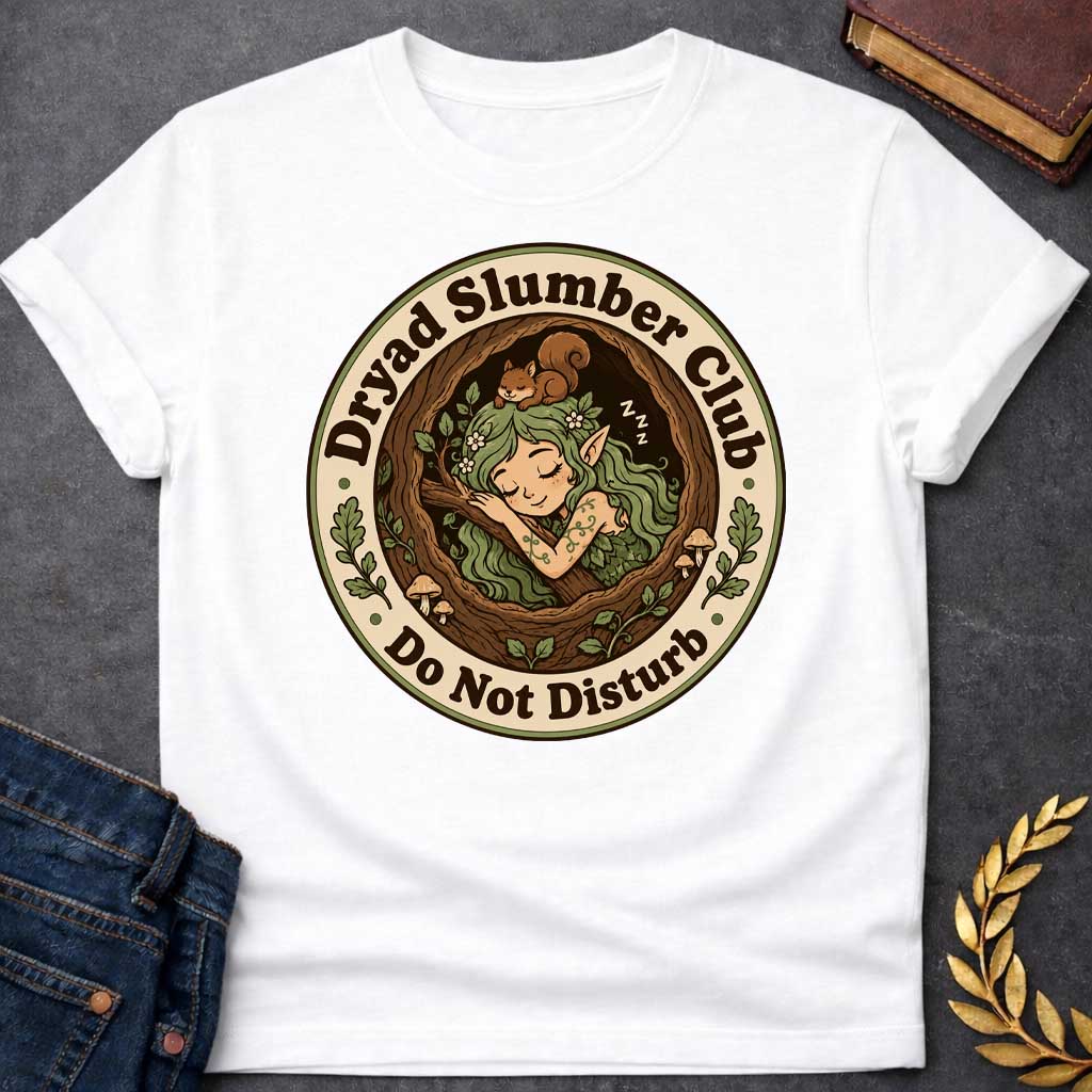 White T-shirt with a cute, cartoon-style t-shirt graphic enclosed in a circular badge. Inside a cozy tree hollow, a little green-haired dryad or forest elf sleeps peacefully next to small mushrooms, while a tiny brown squirrel naps on her head. A cream-colored outer ring features dark brown text that reads "Dryad Slumber Club" along the top and "Do Not Disturb" along the bottom, accented by small green oak leaves