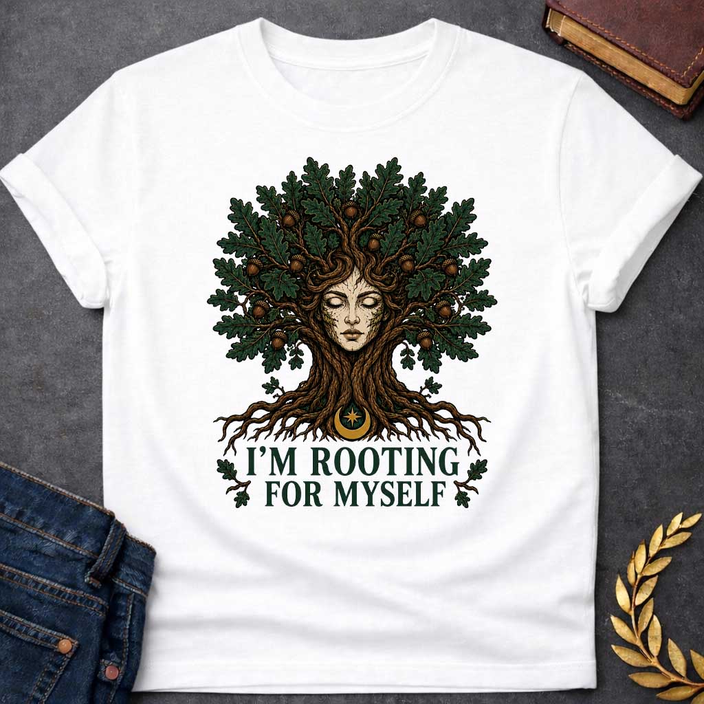 White T-shirt with a cottagecore-style graphic featuring a detailed illustration of an oak tree dryad or nature spirit. Her serene, closed-eyed face forms the center of the tree trunk, with gnarled roots expanding below and a massive canopy of dark green oak leaves and brown acorns spreading above. Underneath the roots, a pun-based tagline reads "I'M ROOTING FOR MYSELF" in a bold, clean green serif font.