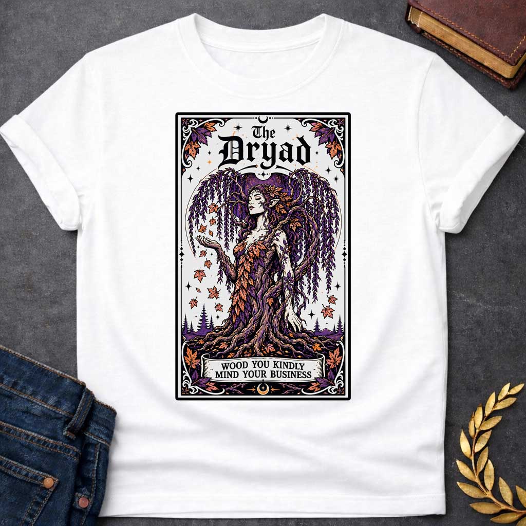 White T-shirt with a mystical, tarot-card style design framed by an ornate border. It features a majestic weeping willow dryad whose gown is woven from purple and orange autumn leaves. At the top, bold gothic text reads "The Dryad." A weathered banner at the base of her rooted trunk features a clever, pun-based tagline that reads: "WOOD YOU KINDLY MIND YOUR BUSINESS" in a clean, black serif font