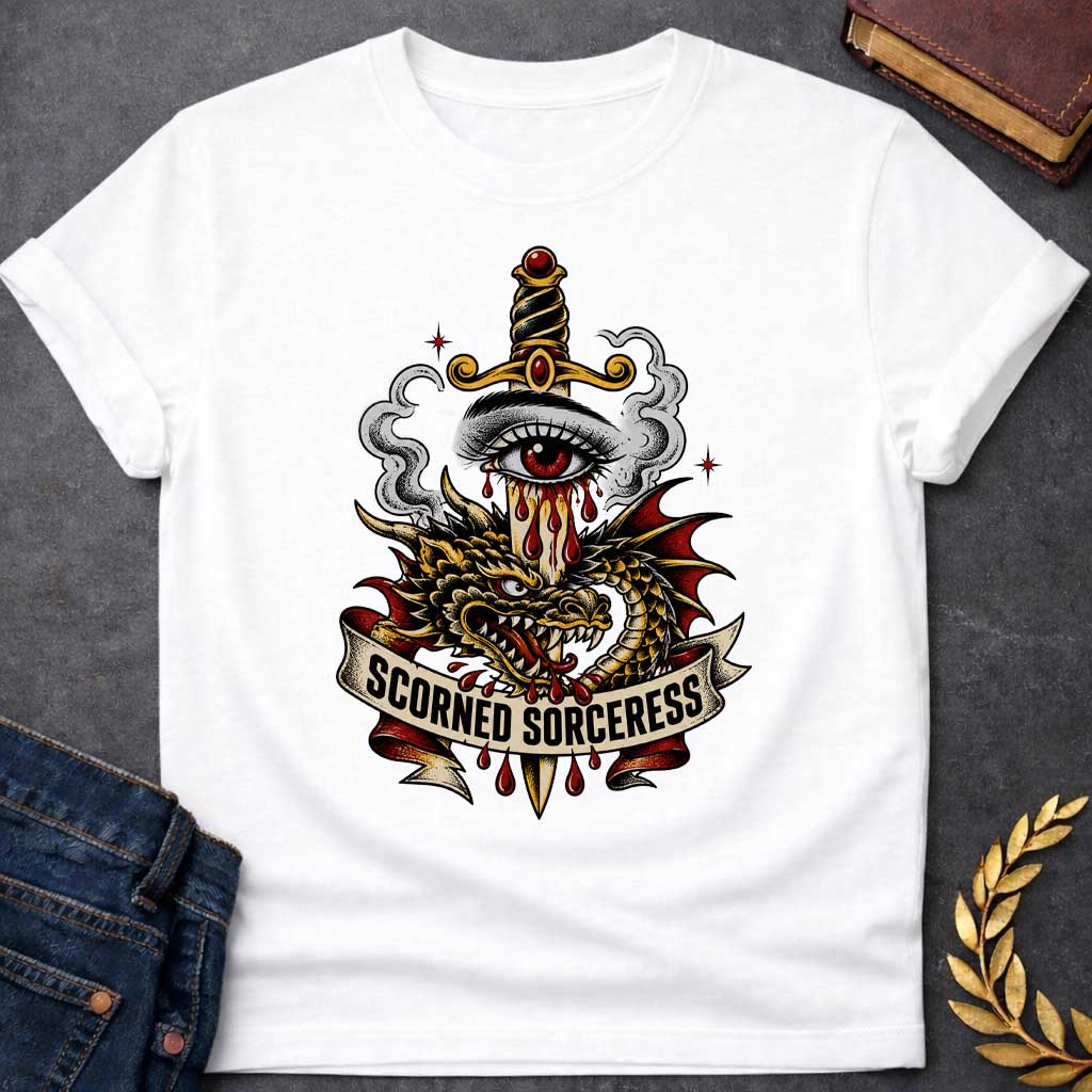 White T-shirt featuring traditional tattoo-style artwork: a gold-hilted dagger with black-and-gold wrapped handle and ruby pommel pierces through a weeping red-irised eye, blood streaming down the blade. A gold and red dragon coils around the dagger, fangs bared, with grey smoke curling behind. A cream ribbon banner below reads "Scorned Sorceress" in bold black lettering, dripping blood, on a white background.