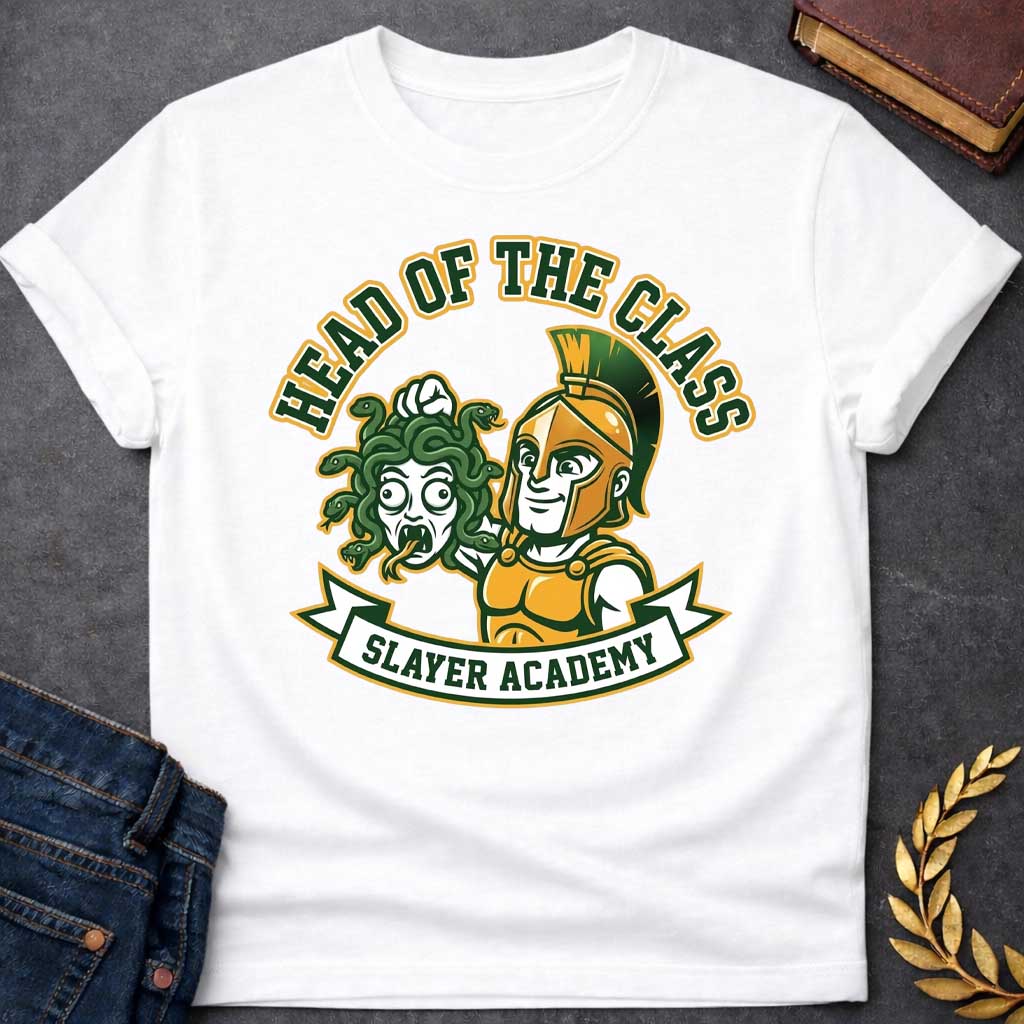 White t-shirt in collegiate mascot style: a smirking gold-helmeted Greek warrior holds up Medusa's shocked, snarling severed head by her green snake hair. Bold green and gold text arcs above the design, with "Slayer Academy" on a white ribbon banner below. Playful varsity-logo aesthetic on a white background.