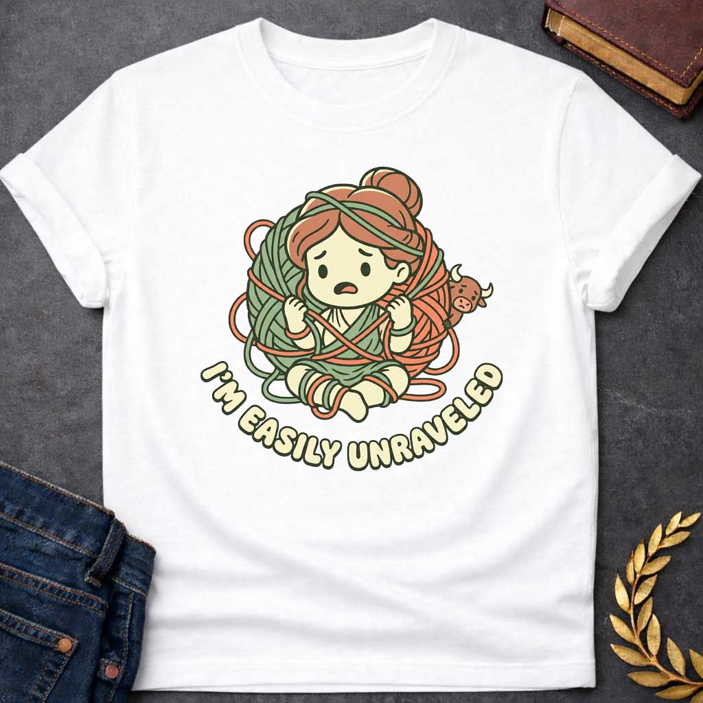 White T-shirt with A cute chibi-style t-shirt graphic. A worried cartoon Ariadne sits tangled inside a giant ball of green and orange yarn, while a tiny, peekaboo Minotaur looks on from behind. Below the illustration, the punny tagline "I'M EASILY UNRAVELED" arcs gracefully in a bubbly, retro font. The design features a charming pastel color palette of muted sage green, terracotta orange, and cream.